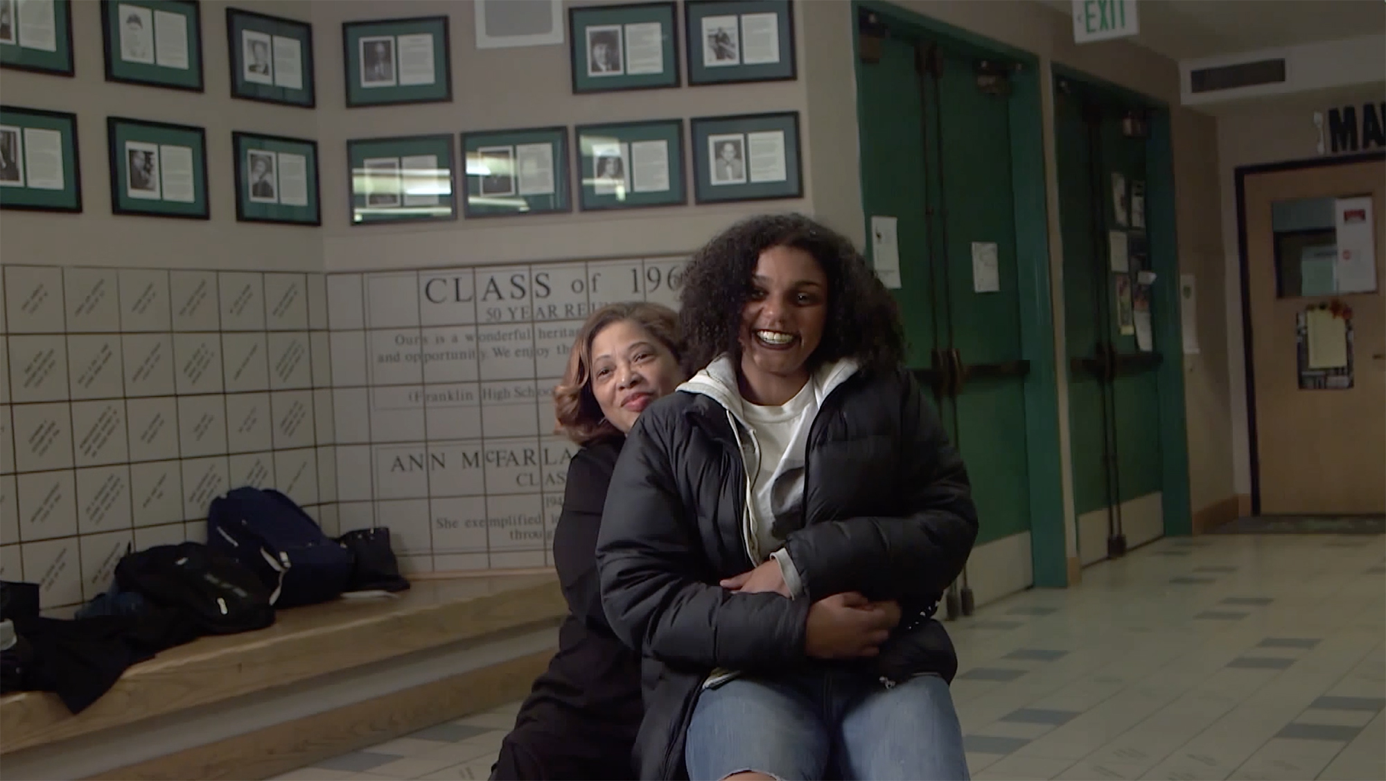 Woman, seated, with younger woman on her lap, in the hallway of a school.