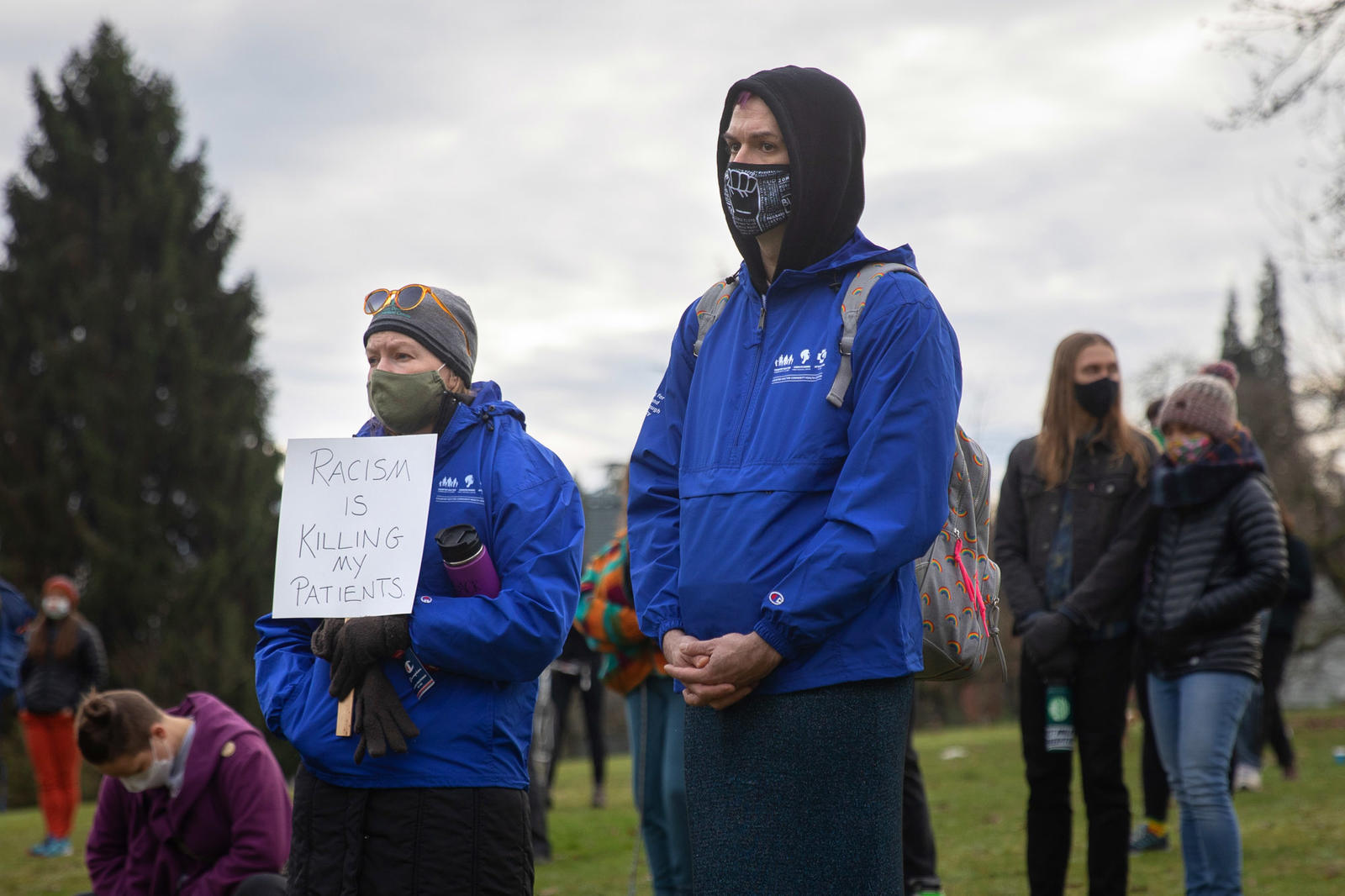 Protesters standing in a park. One holds a sign reading "Racism is killing my patients."