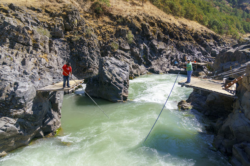 Two fishers stand on platforms on either side of a river holding fishing nets