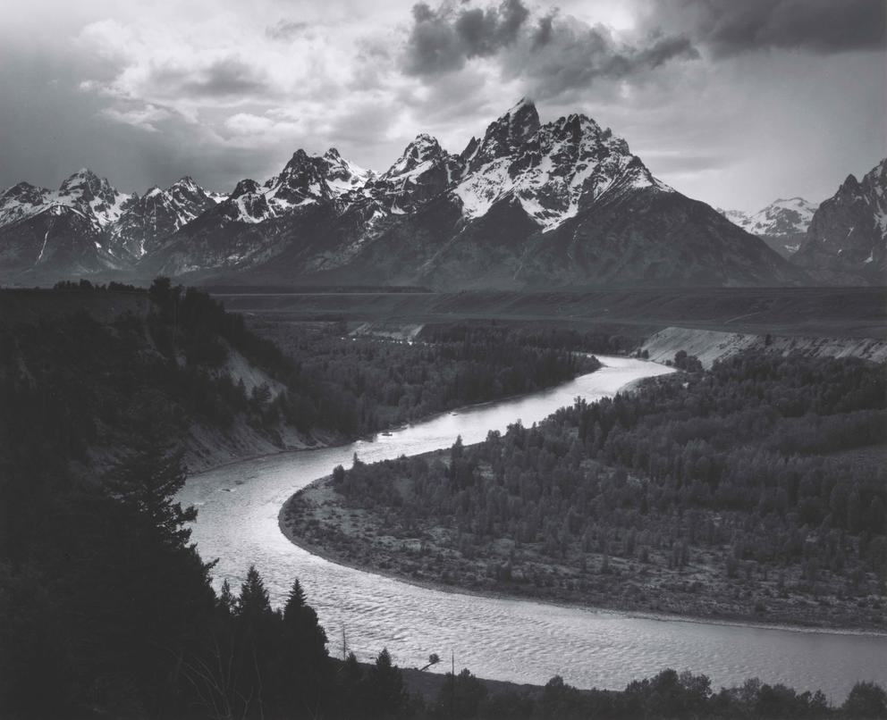 black and white photo of a river with snow-capped mountains int he distance, clouds overhead