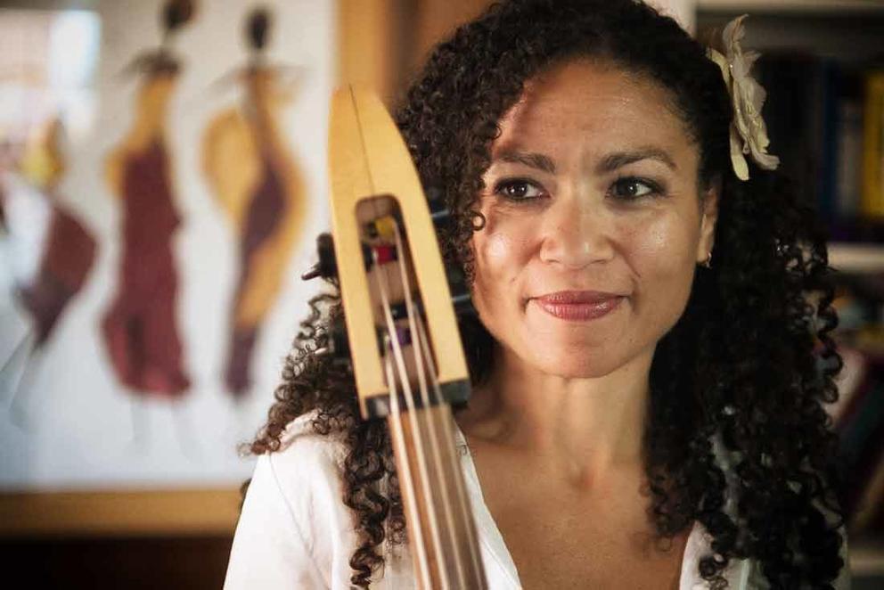 a smiling woman with curly brown hair shown from the shoulders up, with the neck of a cello visible