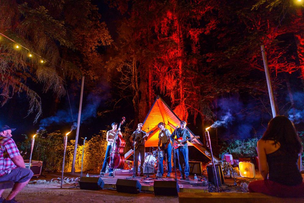 Band plays in the dark, in front of a pyramidal structure that lits up in an orange glow. behind the band is a dark forest, trees rising up tall