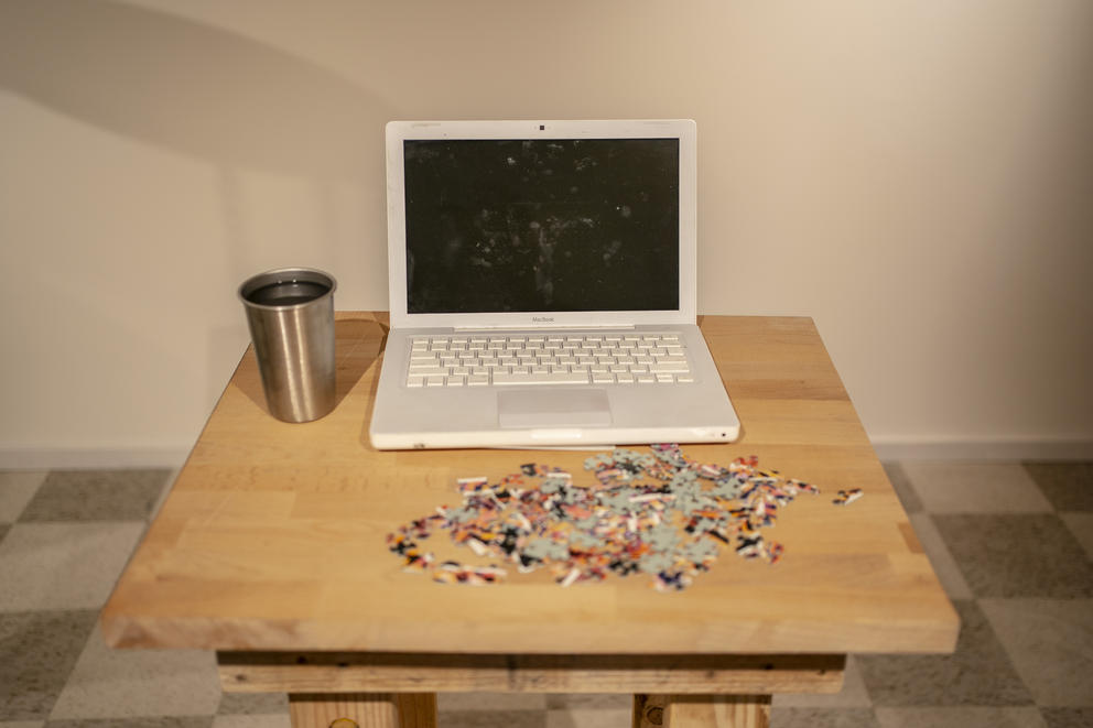 A white laptop sits on a small wood table with a silver cup to its left and a pile of puzzle pieces on the table in front of it