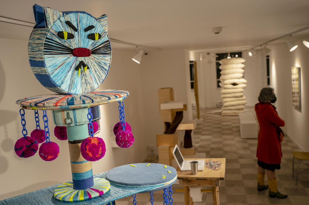 A wide shot of a colorful cat tower sculpture made of yarn in the foreground, a woman in the background looks at other sculptures