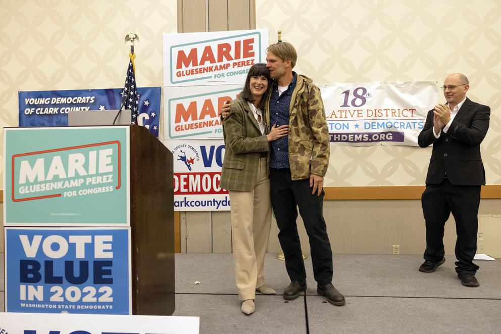 two people hug on stage a man stands at right clapping