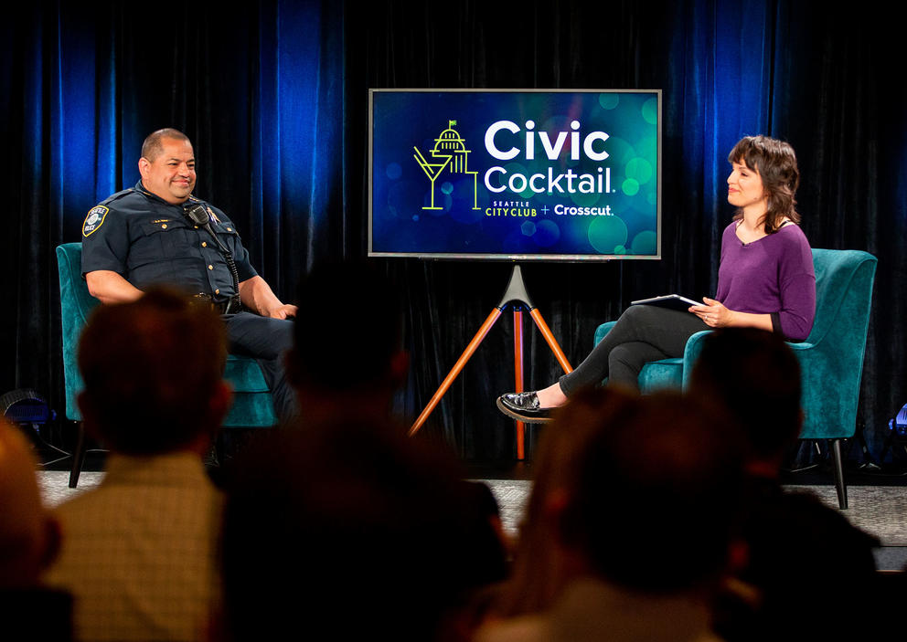 Two people sit in armchairs on a stage in front of a crowd