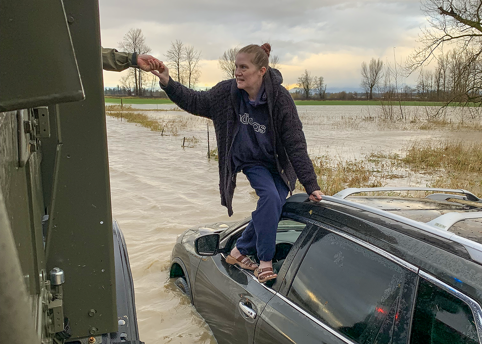 A woman balances on a car door's window well and grasps a hand reaching for her. The car is mostly submerged in floodwater