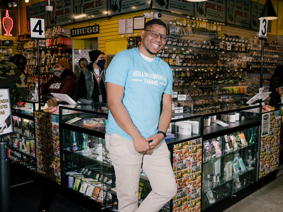 a man in an aqua colored shirt and light colored pants leans back against a glass display case full of marijuana products