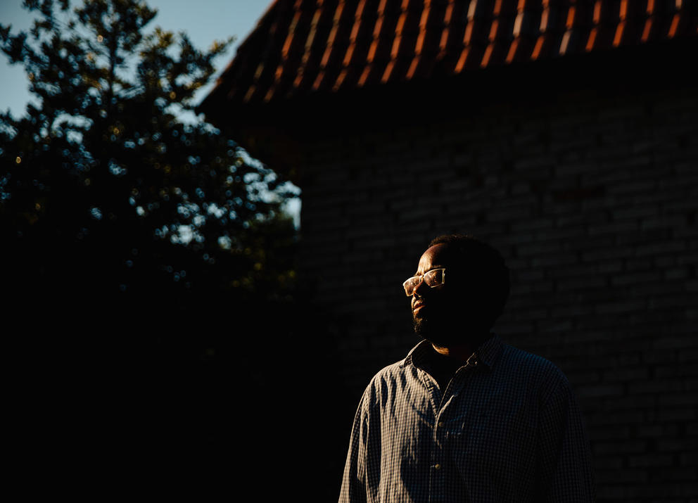 A man with glasses stands in shadow with a sliver of light hitting his left side