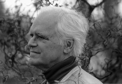 a man with white hair and wearing a dark turtleneck looks into the woods