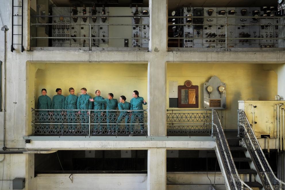 photo of a large industrial space, with dancers lined up in blue coveralls