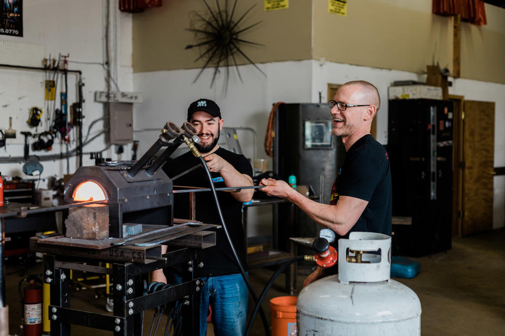 Two people are seen laughing and working in a blacksmithing studio