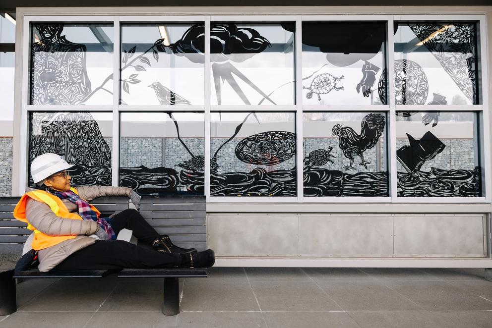 a woman wearing a hard hat and safety vest sits on a bench near a glass windscreen adorned with art