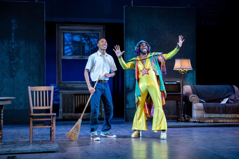 a photo of two people performing on stage, one a young boy in street clothes sweeping, the other in a psychedelic yellow jumpsuit