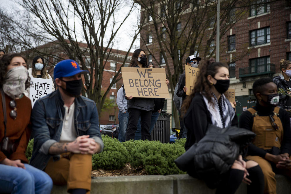 A group of demonstrators standing in a park. One holds a cardboard sign reading "We belong here"