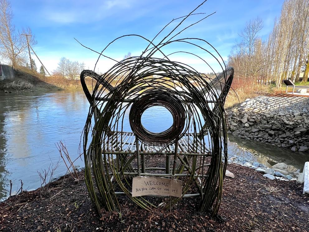 photo of a bench at the edge of a river, made from bent willow branches