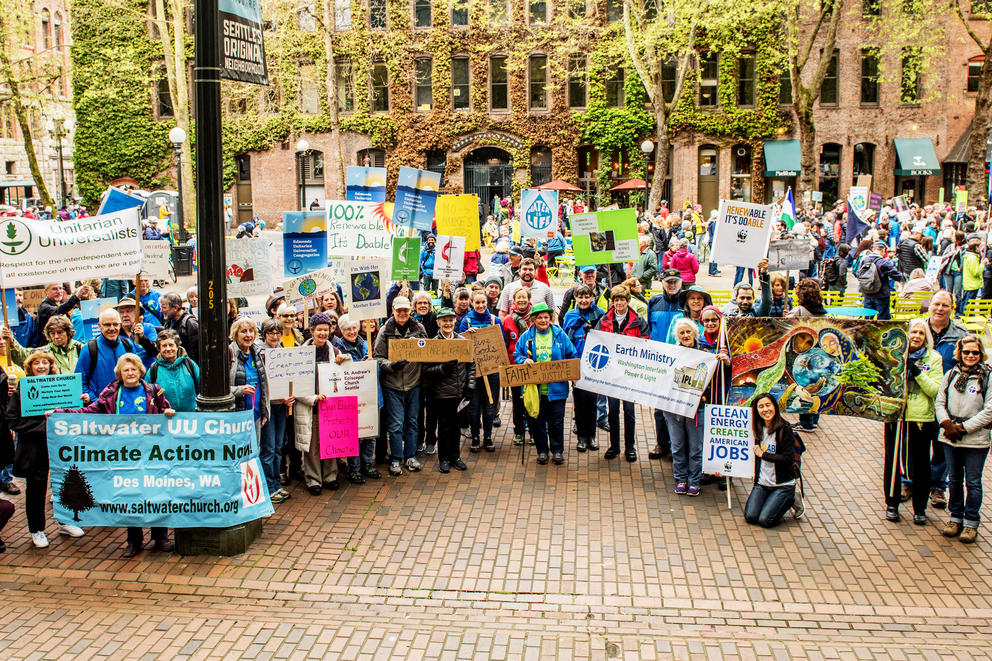 A large crowd of protesters hold signs representing their faith groups, including Unitarian Universalists, Saltwater UU Church, and Earth Ministry, in addition to posters reading "Clean energy creates American jobs" and "Faith = climate justice"