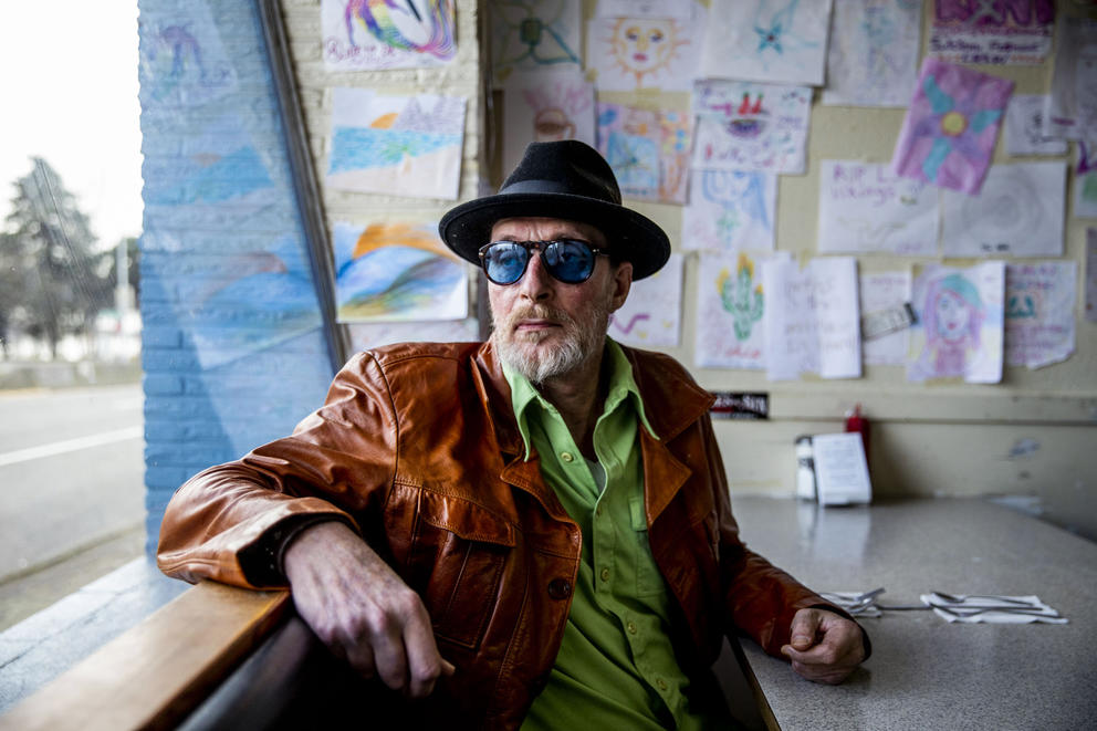 a man wearing a jacket and hat looks out a window, leaning on the sill