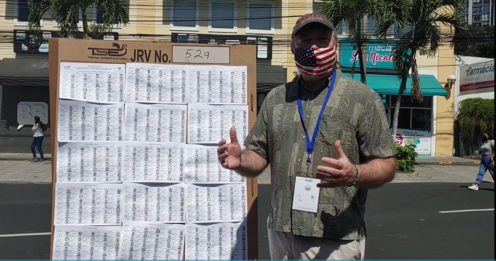 Doug Ericksen stands in front of a poster talking about the voting process in El Salvador, with palm trees and buildings behind him