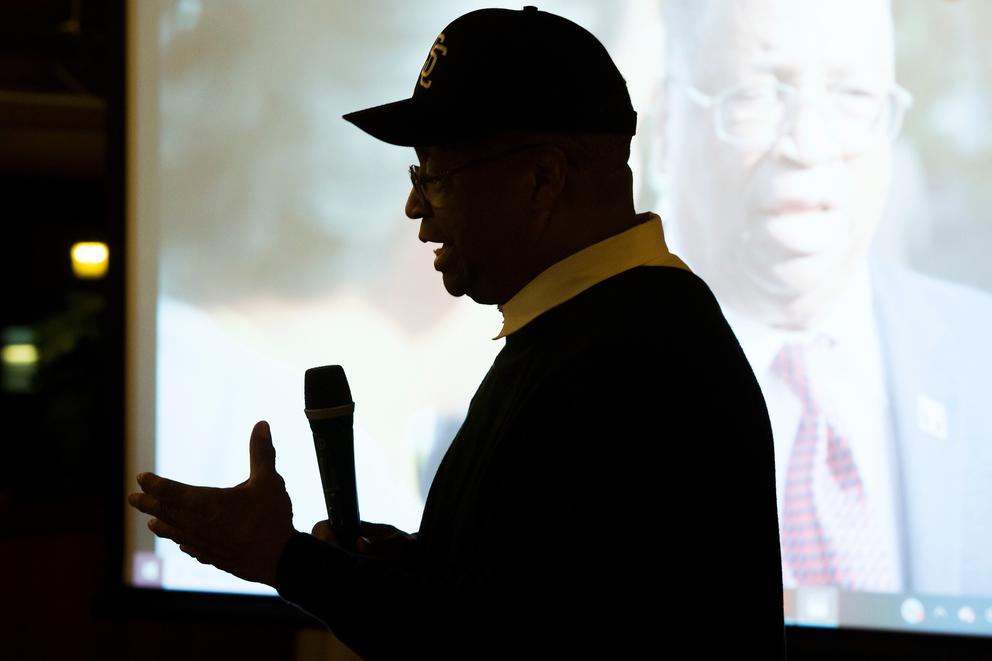 Larry Gossett holds a microphone and speaks to a crowd, with a campaign video playing behind him.