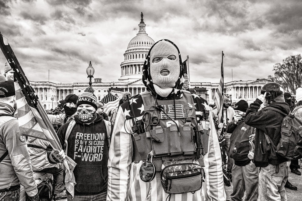 Black and white photo of person with a mask and protective gear in front of a crowd and the capitol building in Washington, DC