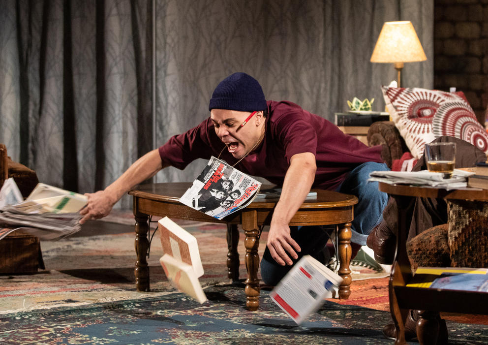 Person on a stage with lamp in the background throws magazines and books off a table, leaning over and with arms outstretched. They are wearing a bordeaux sweater.