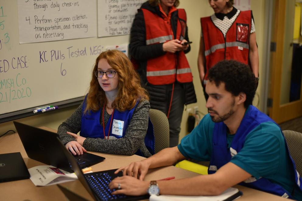 A man and woman in vests work at computers in front of a whiteboard