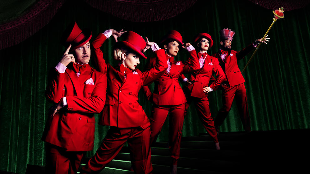 Six figures dressed in bright red clothing in a dark room, stand on stairs ascending from left to right