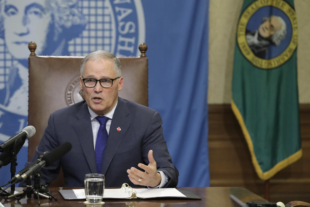 Gov. Jay Inslee sits with the state seal of Washington and state flag behind him.