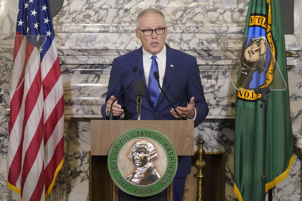 Man in suit at podium with American flag and state of Washington flag to the sides of him. Background is marble, podium has gold state seal