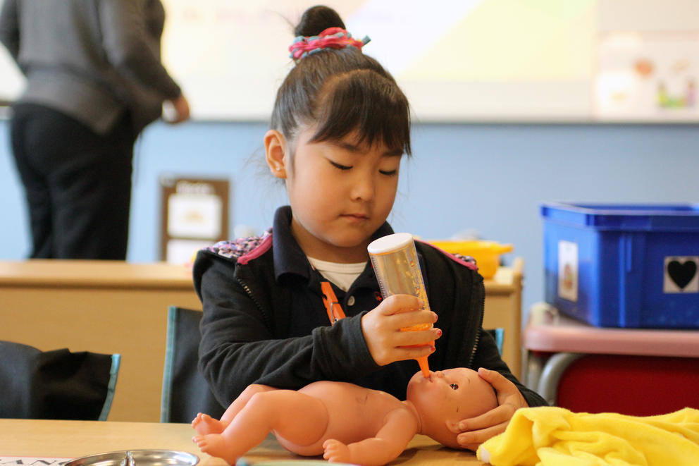 A young girl sits at a desk with a baby bottle and doll