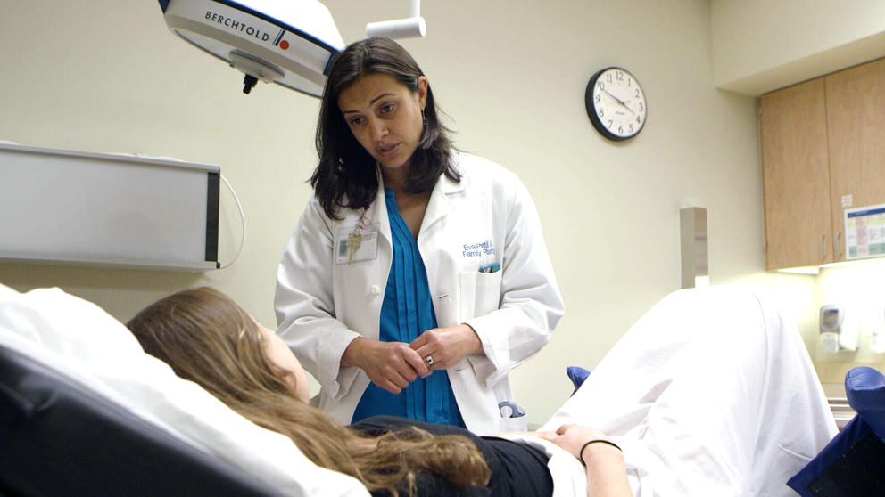 a woman doctor with dark brown hair leans over another woman lying back in a doctor's office