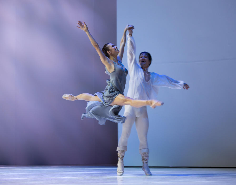 two ballet dancers performing on stage, the woman leaping in the splits, man supporting