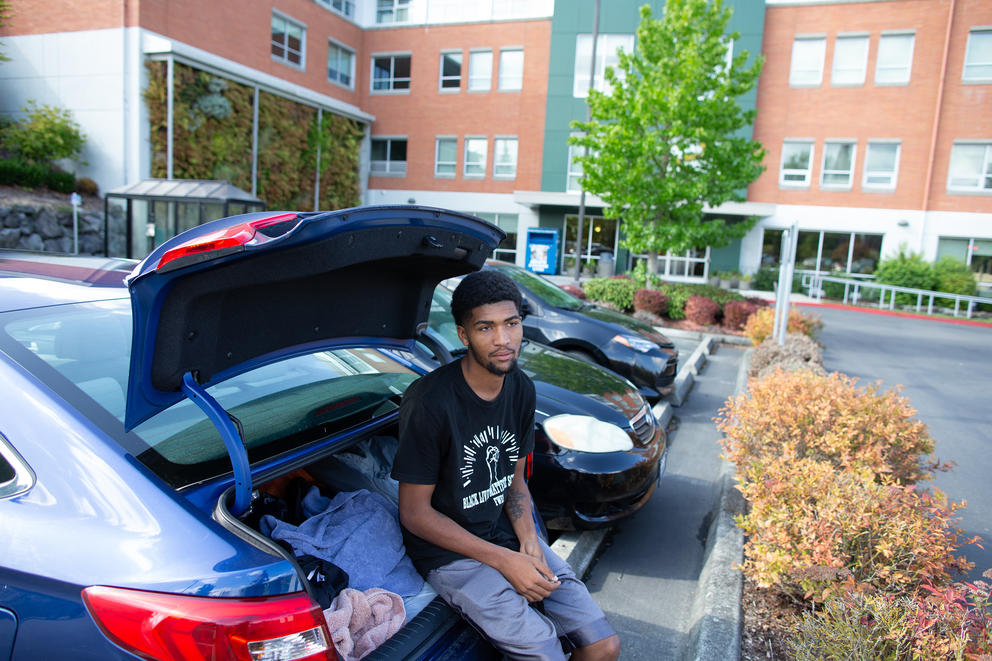 A young man sits on the bumper of a car in a parking lot.