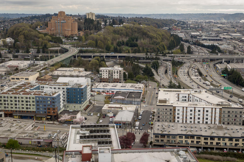 Image of city buildings on a cloudy day. Many highways run between them. In the background, a copse of trees is completely encircled by roads. 