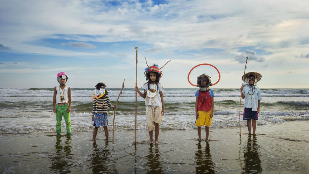 a row of children stands at the edge of the ocean, decked out in detritus