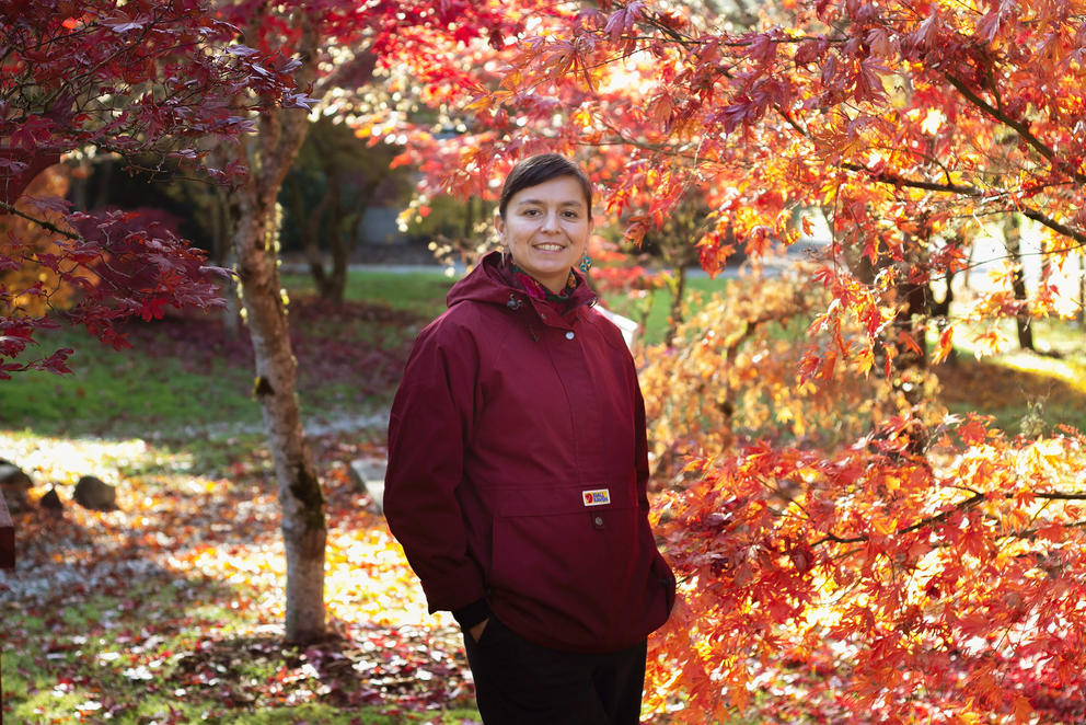 Person smiles in front of fall foliage, she is wearing a wine-red coat