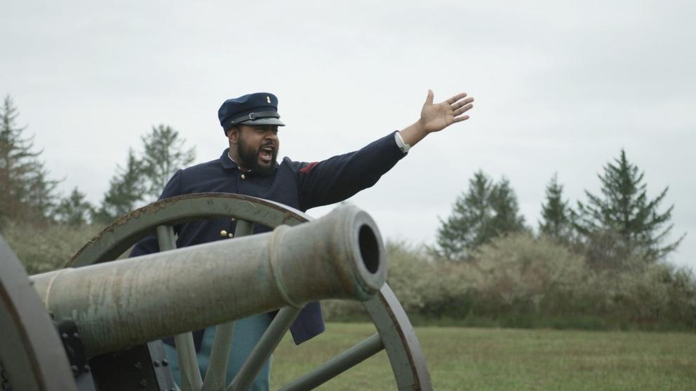 A movie still featuring a Black man in military dress shouting next to a cannon