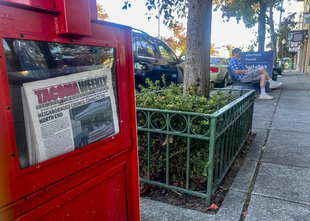 A red newspaper box with copies of the Tacoma Weekly, with a man sitting on a bench in the background