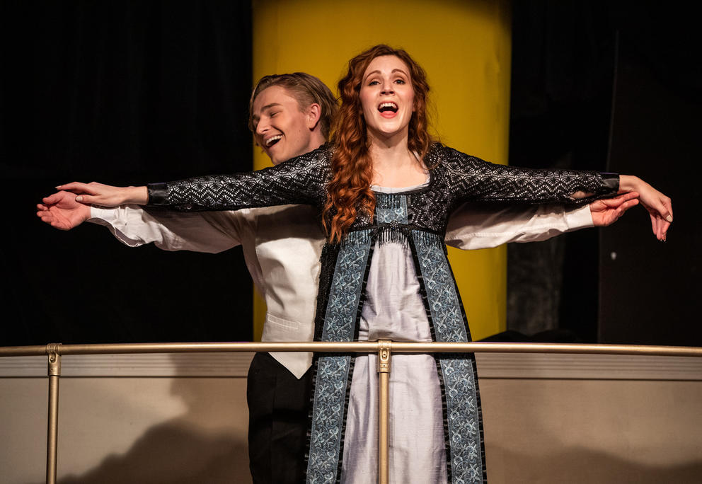 Woman in a white and blue dress stands with arms stretched out, a man standing behind her. They both appear to be singing.