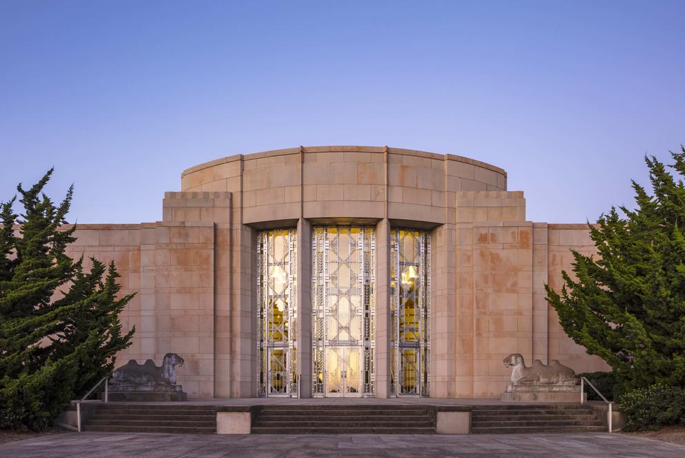 Art deco building in front of a blue sky, framed by two large green bushes. The aluminum front doors reflect the light of the sunlight. The building front looks almost rosé.