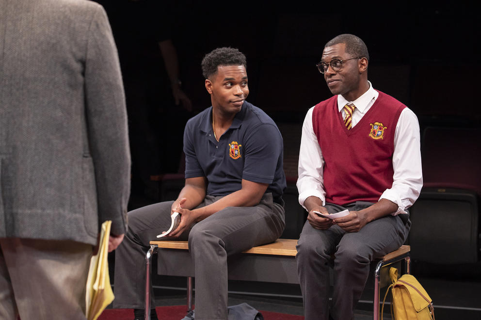 Two actors look at each other as they sit on a bench