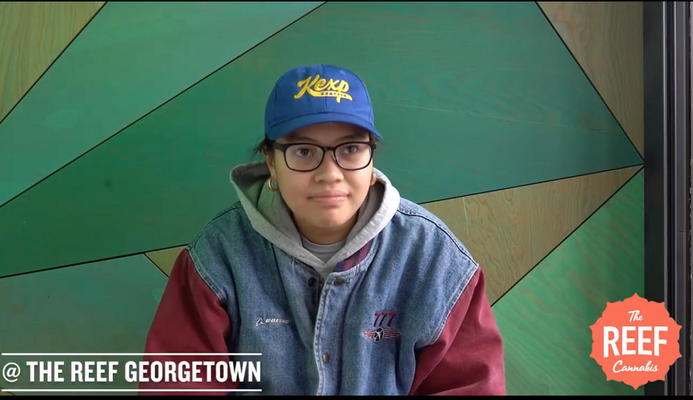 screen grab of a woman wearing a large coat, glasses and baseball cap, with the logo for The Reef on screen