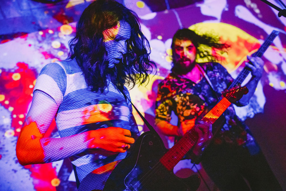 Two people play on guitars as colorful lights hit them from above, creating a psychedelic, kaleidoscopic effect