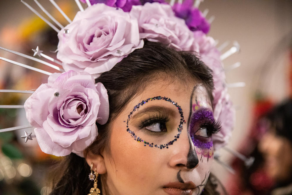 A close up of a woman's face, she is wearing Day of the Dead face paint and a crown of paper flowers