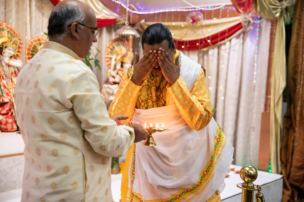 A man holds out an oil lamp with lit flames beside another man in yellow who holds his hands to his face above the flame