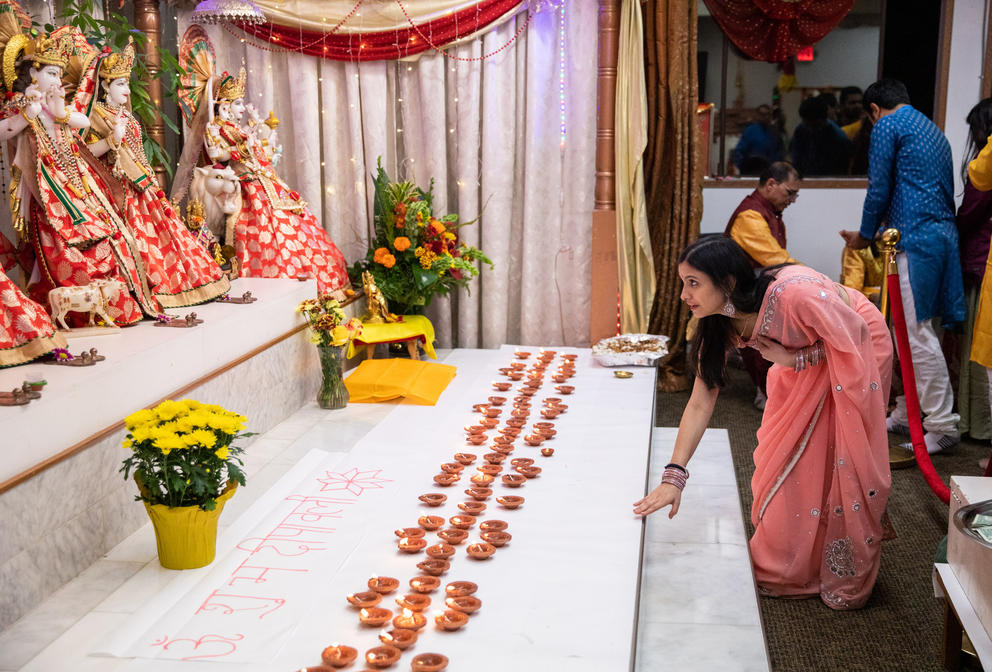 A woman in a pink dress bends down before a stage lined with candles and deity figures.