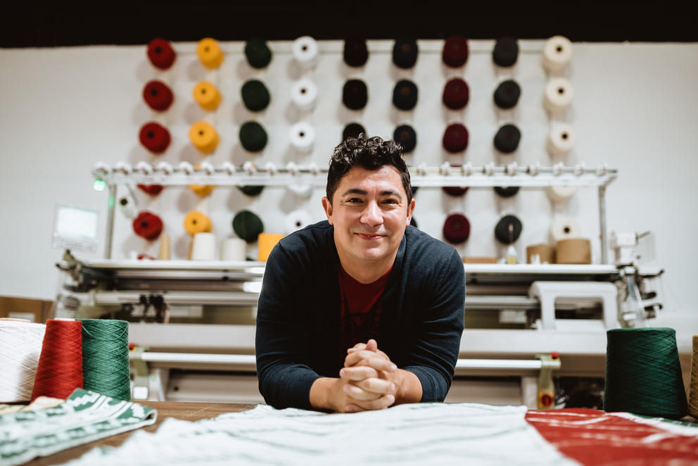 Person in front of knitting machine, smiling. Colorful spools of yarn on the back wall.