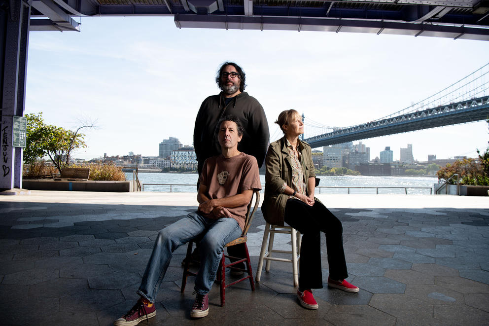 One man and a woman sit next to each other on chairs, while another man stands between them