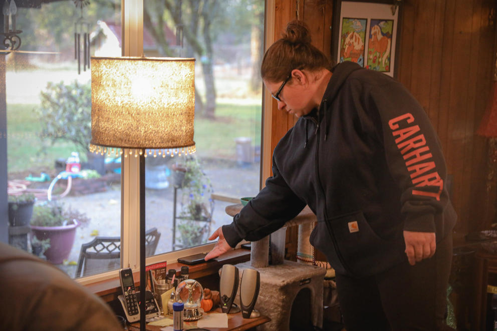 A woman reaches out to set a phone down on a windowsill inside a house next to a table lamp